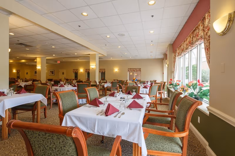 Spacious dining room with neatly set tables, green-upholstered chairs, folded burgundy napkins, and large windows with plants.