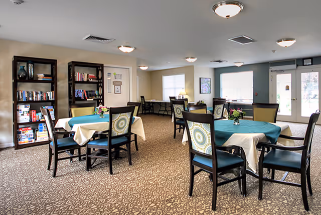 A well-lit common room with several tables covered with white and teal tablecloths, each surrounded by chairs with patterned cushions. There are bookshelves filled with books and board games along one wall, a desk with a lamp near a window, and a glass door leading outside. The room has a patterned carpet and ceiling lights.