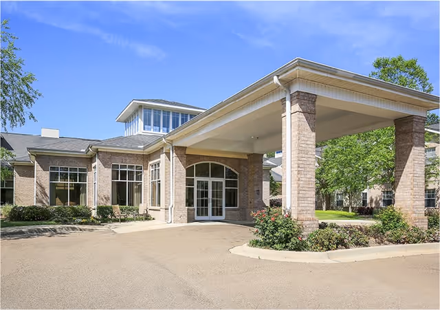 Front entrance of a brick senior living building with a covered porte-cochere, large windows, and landscaping under a blue sky.