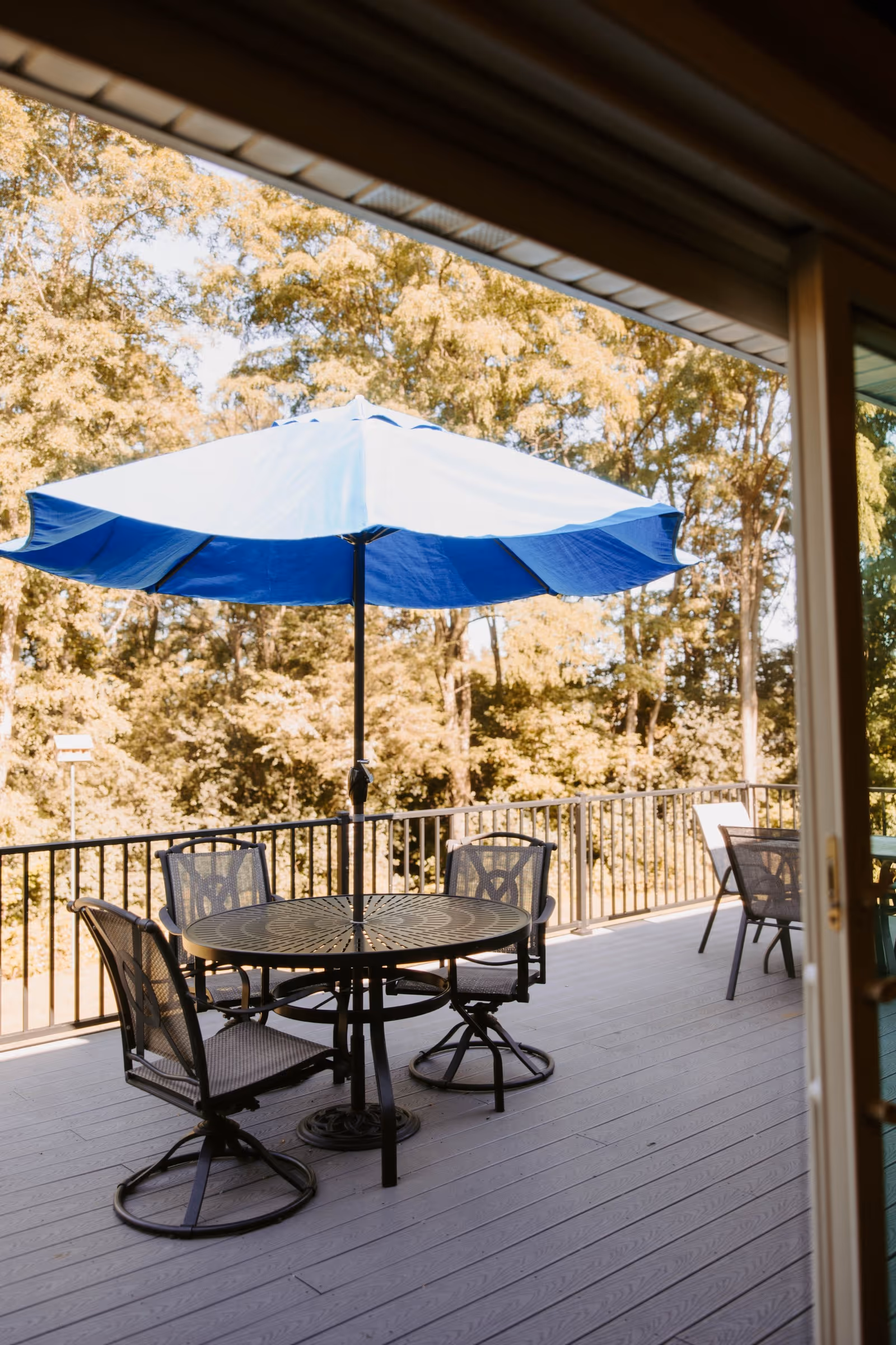 Outdoor patio area with a round metal table and four matching chairs under a large blue umbrella, surrounded by a railing and trees in the background.