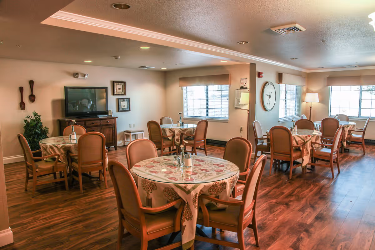 A dining room with multiple round tables covered with floral tablecloths, each surrounded by wooden chairs with cushioned seats. The room has wood flooring, large windows with blinds, a TV on a wooden cabinet, wall decorations, and a floor lamp in the corner providing warm lighting.