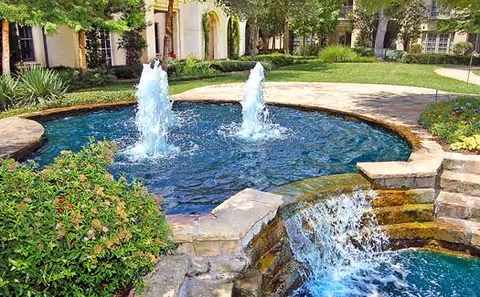 Outdoor garden area with a circular water fountain featuring two vertical water jets, surrounded by stone edging and greenery, with a small waterfall flowing into a lower pool. In the background, there are trees, shrubs, and a building with arched windows.