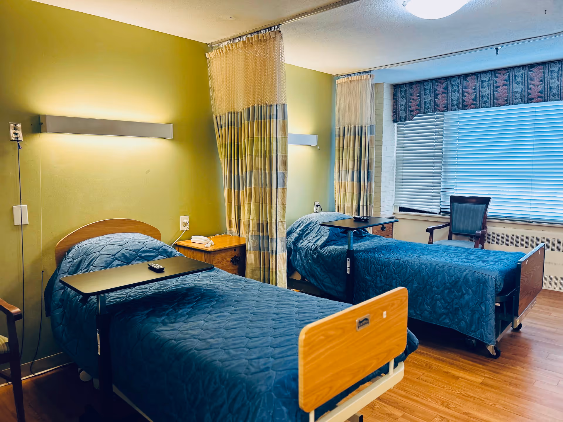 A room with two hospital-style beds separated by a curtain. Each bed has a blue quilted blanket and an overbed table. There is a wooden nightstand between the beds with a telephone on it. The walls are painted green, and there is a window with blinds and a patterned valance. A chair is positioned near the window.