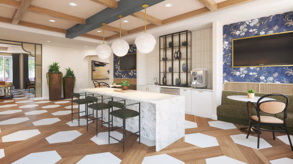 A modern common area with a large marble island countertop surrounded by four black chairs with green cushions. Three round pendant lights hang above the island. The floor features a geometric pattern with wood and white hexagonal tiles. In the background, there is a built-in cabinet with a coffee machine and decorative shelves, a green cushioned bench with a round table and chairs, and a wall-mounted flat-screen TV. The ceiling has wooden beams and recessed lighting.