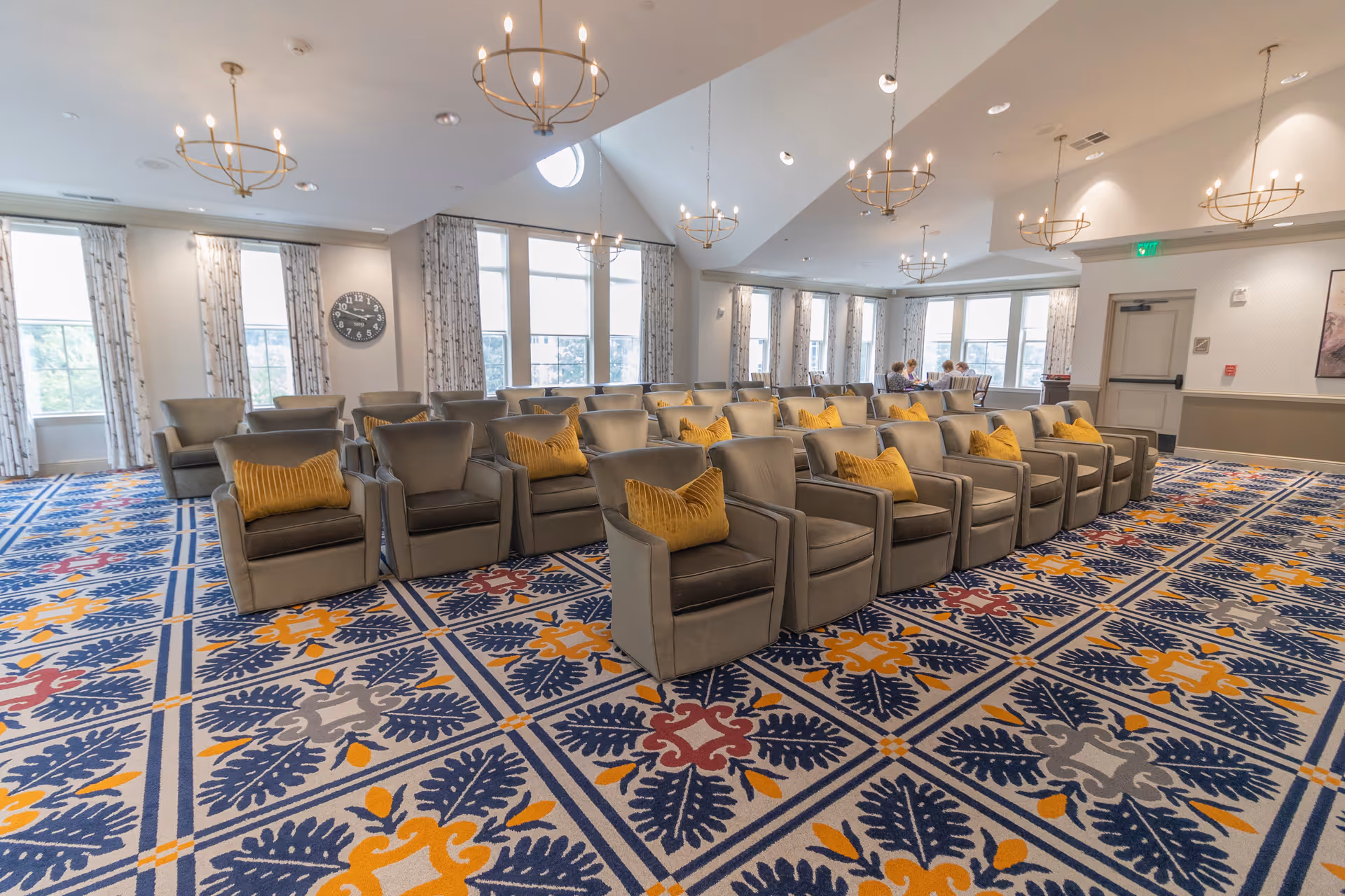 Spacious senior living common room with rows of gray armchairs with yellow pillows, patterned carpet, large windows and chandeliers.