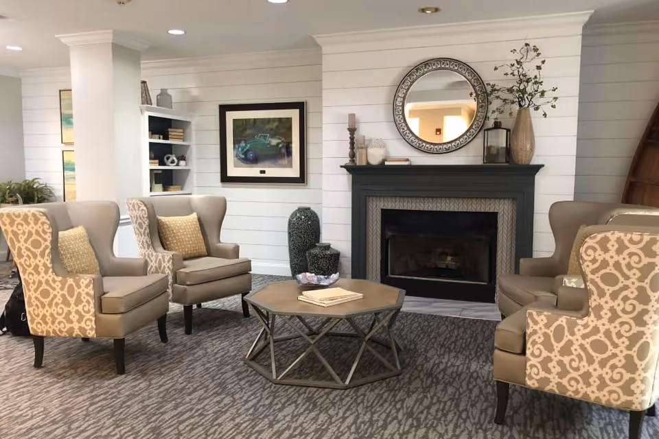 A cozy living room area with four beige armchairs arranged around a hexagonal wooden coffee table. Behind the table is a dark fireplace with decorative items on the mantel, including a round mirror, candles, and a vase with branches. The walls are white with horizontal paneling, and there is a framed picture and a built-in bookshelf in the background.