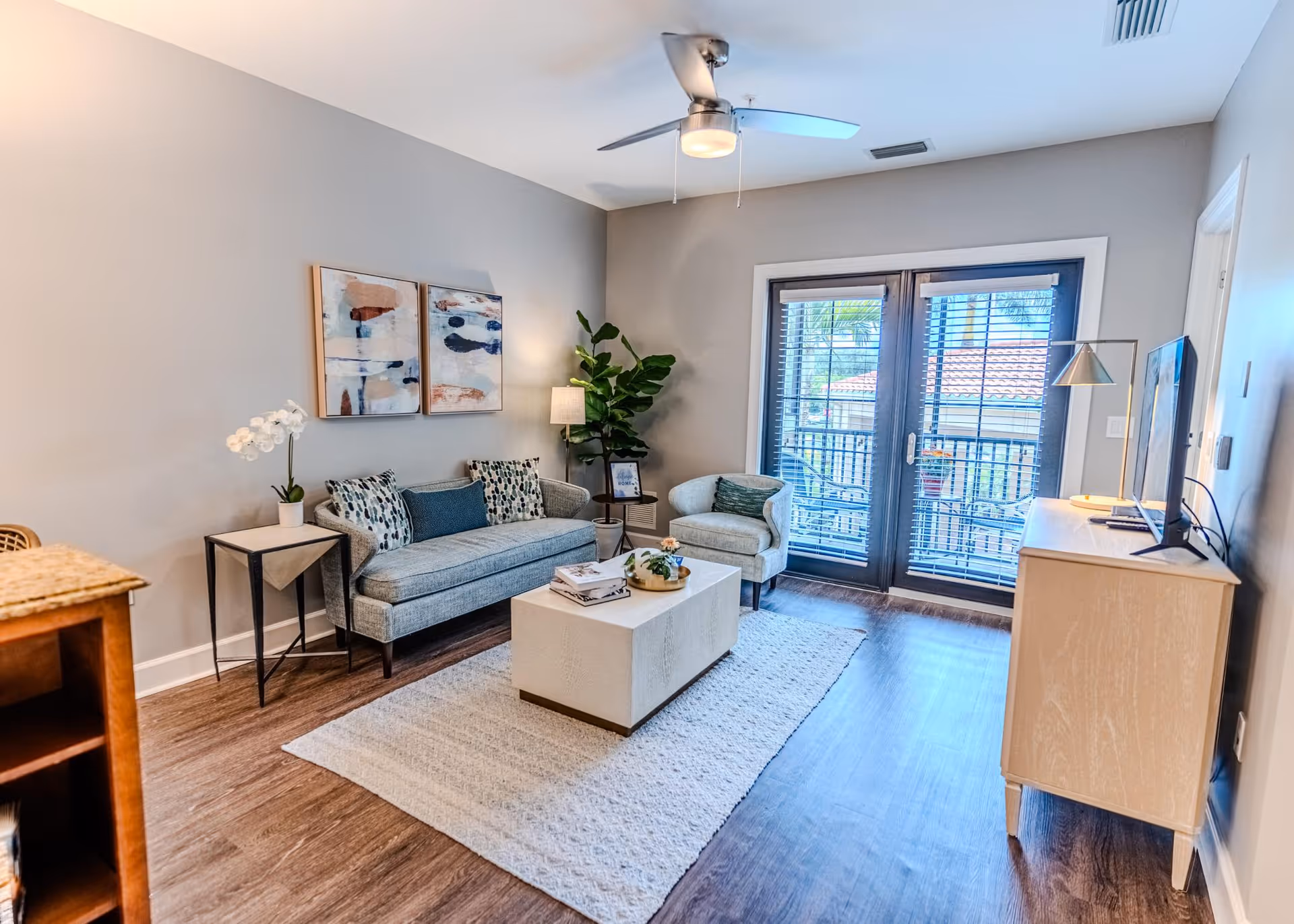 A bright living room with a gray sofa, an armchair, a white coffee table on a light rug, and a wooden TV stand with a flat-screen TV. There are two abstract paintings on the wall, a potted plant, a floor lamp, and large glass doors leading to a balcony with outdoor seating.