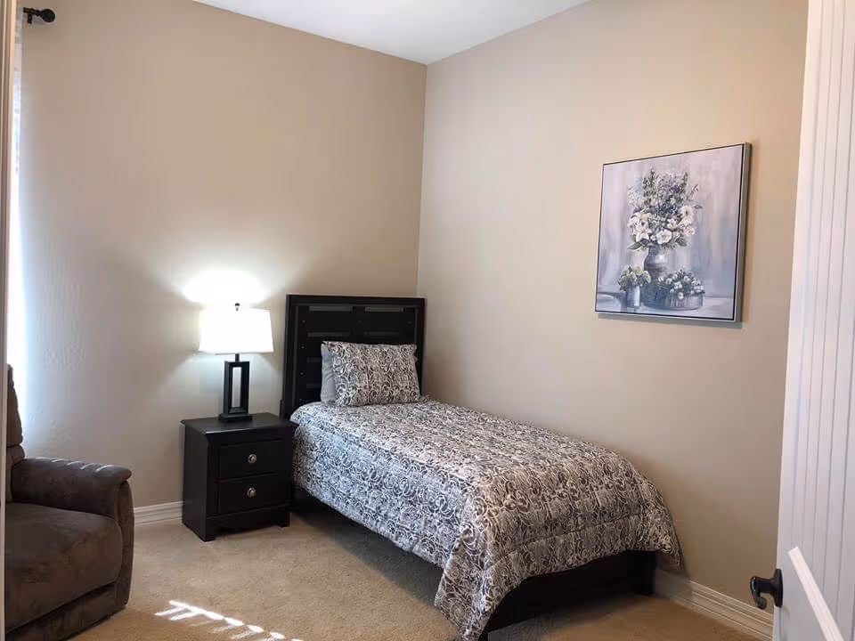 A small bedroom with a single bed covered in a patterned gray and white bedspread and matching pillow. Next to the bed is a dark wooden nightstand with two drawers and a modern table lamp with a white shade. A brown upholstered armchair is partially visible on the left side. The walls are beige, and a framed painting of a floral arrangement hangs above the bed. The room has beige carpet and a white door is partially open on the right side.