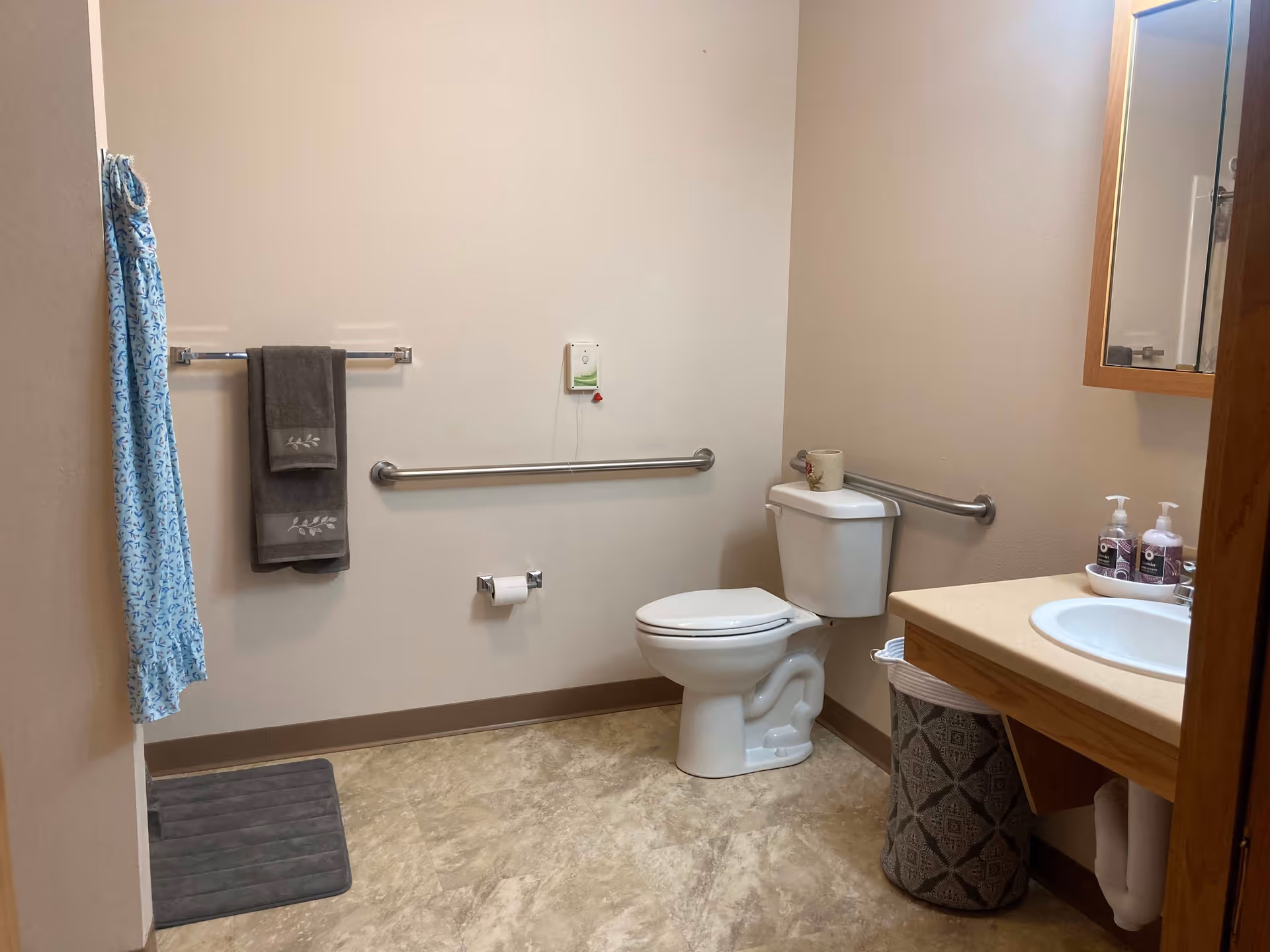 A clean and accessible bathroom with a white toilet, grab bars on the walls, a towel rack with two gray towels, a blue patterned shower curtain, a gray bath mat, a sink with a wooden cabinet and mirror above it, and a laundry basket underneath the sink.