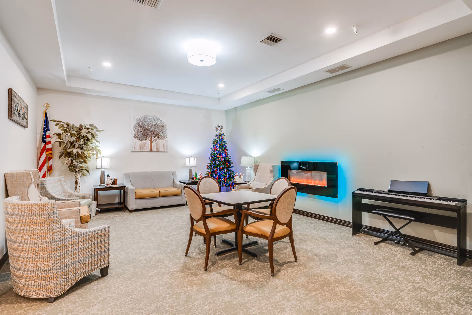A bright common lounge with chairs and a sofa around a table, an electric wall fireplace, keyboard, and a decorated Christmas tree.