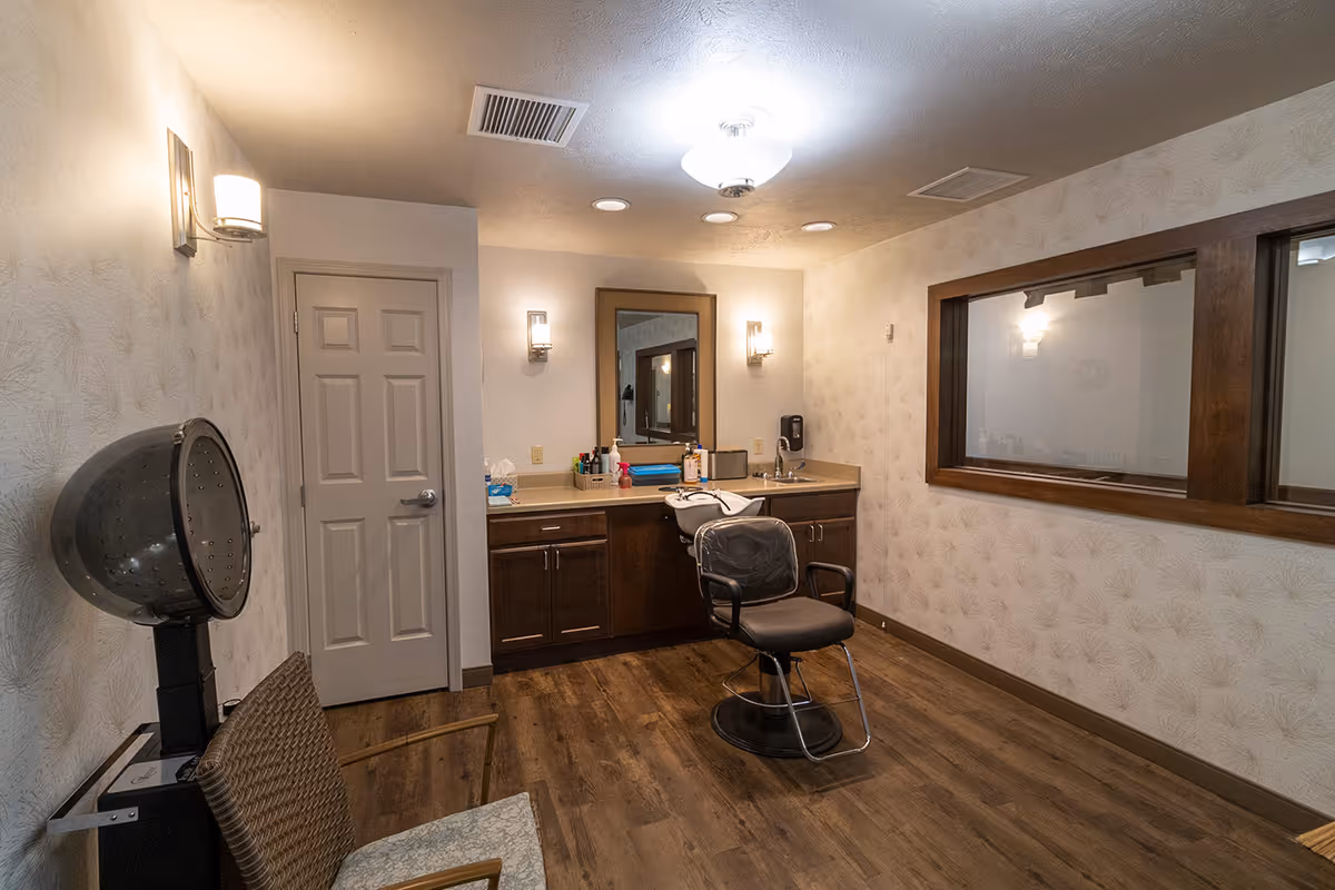 Interior view of a hair salon area in an assisted living facility featuring a hair drying chair, a styling chair in front of a counter with a sink, mirror, and various hair care products, with wooden flooring and soft lighting.