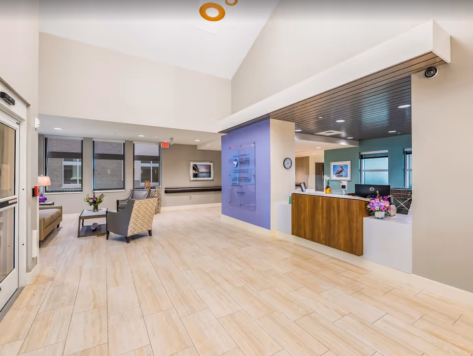 A bright and spacious reception area of a senior living facility with a wooden reception desk on the right, decorated with flowers. To the left, there is a seating area with a sofa, two armchairs, a coffee table with a flower vase, and large windows letting in natural light. The floor is tiled with light-colored tiles, and the walls are painted in neutral tones with some artwork displayed.