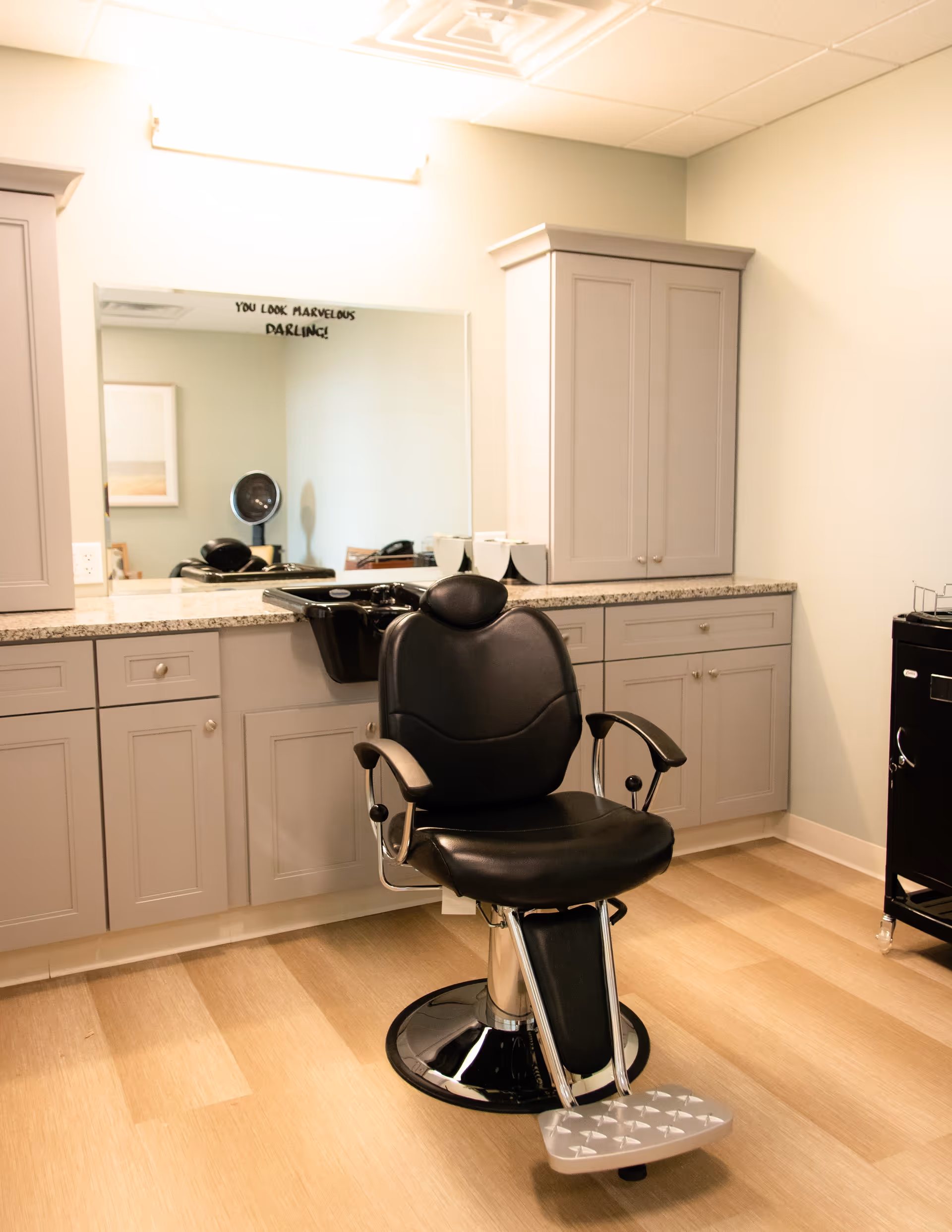 A black barber chair in a clean salon station with cabinets, a countertop and a mirror with a backwash sink.