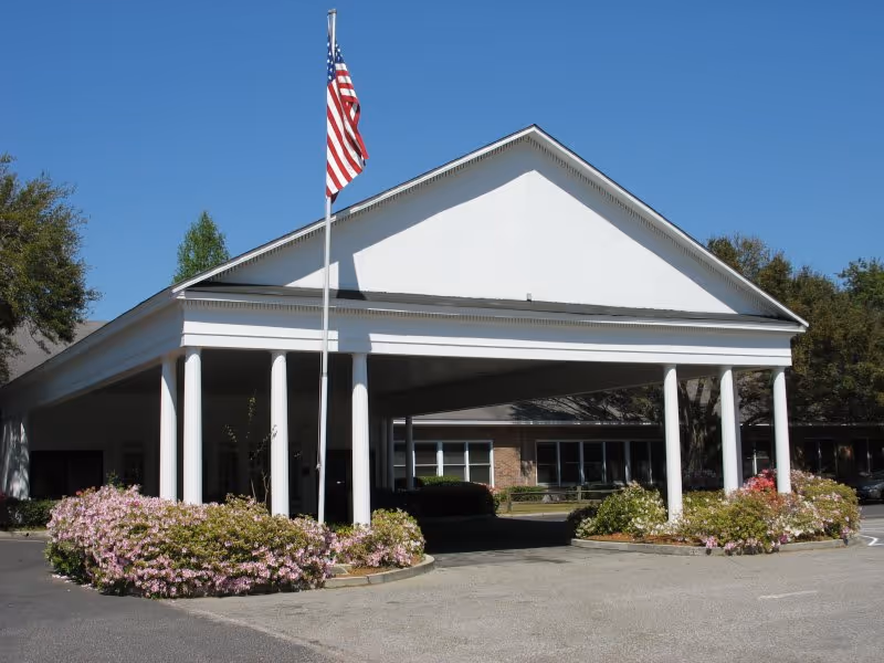 Front exterior view of a building with a large white portico supported by columns, an American flag on a flagpole, and landscaped flower beds with pink flowers in front. The sky is clear and blue.
