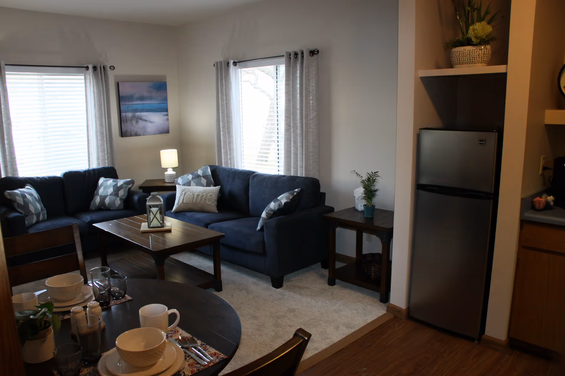 Cozy living room area with two dark blue sofas adorned with patterned and plain pillows, a wooden coffee table with a decorative lantern, and two windows with light curtains. Adjacent to the living room is a small dining table set with white dishes, glasses, and silverware. A stainless steel refrigerator is visible next to a kitchen counter with wooden cabinets. The room has wooden flooring and light-colored walls.
