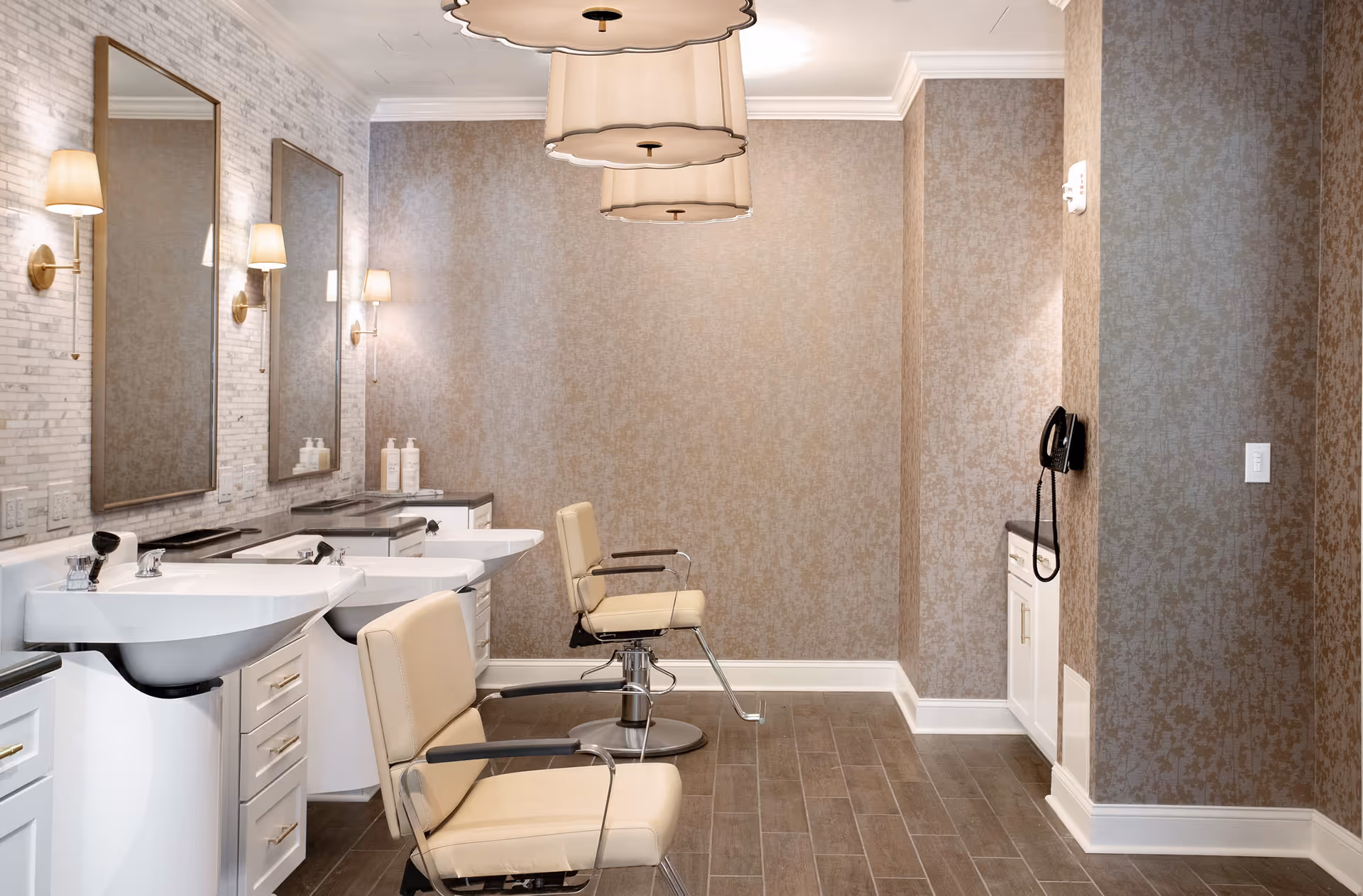 A stylish salon area with two beige salon chairs in front of two sinks and large mirrors mounted on a wall with light-colored textured wallpaper. The room features elegant wall sconces, overhead pendant lights with scalloped edges, and a wall-mounted black telephone. The floor is covered with wood-look tiles.