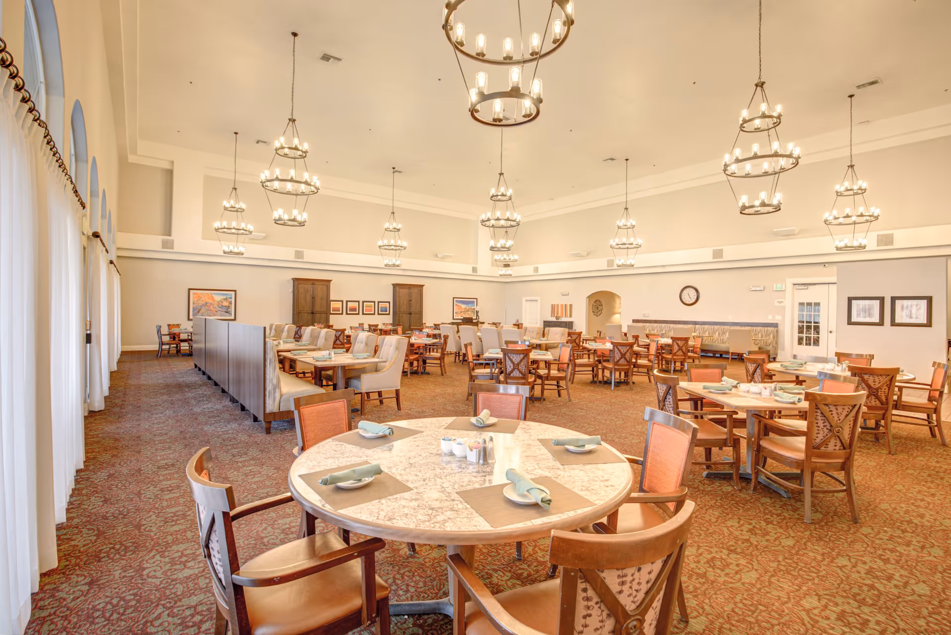 A spacious dining room with multiple round and rectangular tables set with plates, napkins, and utensils. The room features high ceilings with multiple hanging chandeliers, carpeted floors, and large windows with sheer curtains along one side. The walls are decorated with framed artwork and a clock.