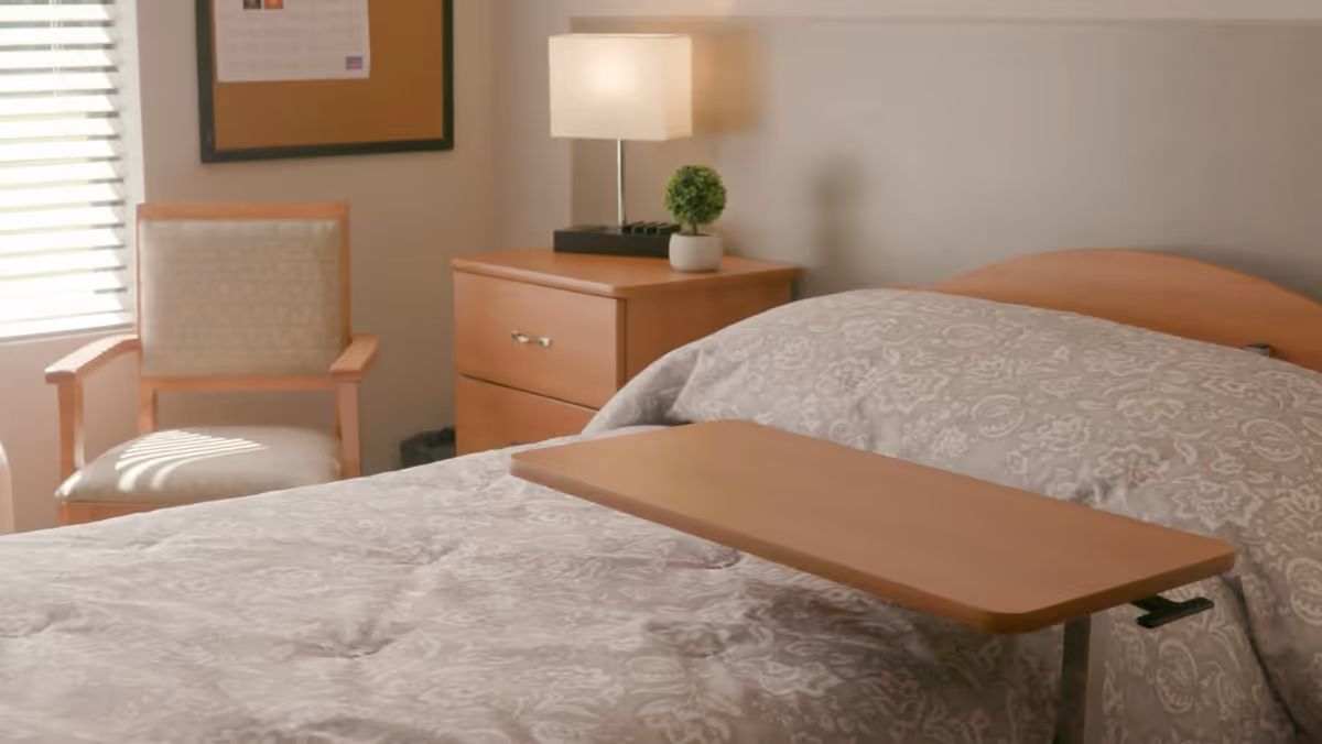 A neatly made bed with a patterned comforter in a nursing center room. A wooden overbed table is positioned on the bed. Next to the bed is a wooden nightstand with a small potted plant and a lamp. A cushioned wooden chair is placed near a window with blinds, and a framed document hangs on the wall.