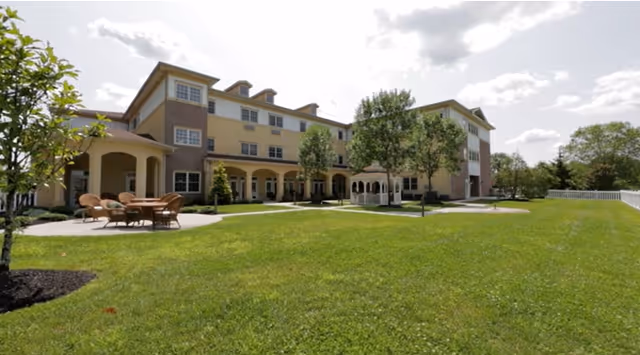 A large, three-story assisted living facility building with a light yellow and beige exterior. In front of the building is a well-maintained green lawn with a white gazebo and outdoor seating area with tables and chairs. Trees and shrubs are planted around the lawn, and the sky is partly cloudy.