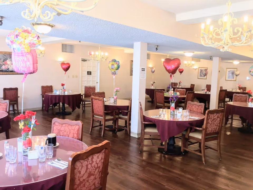 Dining room with multiple round tables covered with maroon tablecloths, each set with glasses, utensils, and floral centerpieces. Heart-shaped and flower-shaped balloons with Mother's Day messages are tied to some tables. The room has wooden flooring, chandeliers, and framed artwork on the walls.