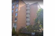 Exterior view of a multi-story brick building with several windows and a stairwell section in the center. There are trees and shrubs in front of the building, and a small sign is visible near the entrance.