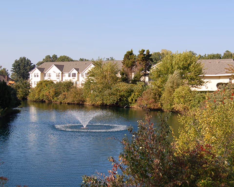 A serene outdoor scene featuring a pond with a water fountain in the center, surrounded by lush green trees and bushes. In the background, there is a large white residential building with multiple windows and a gray roof under a clear blue sky.