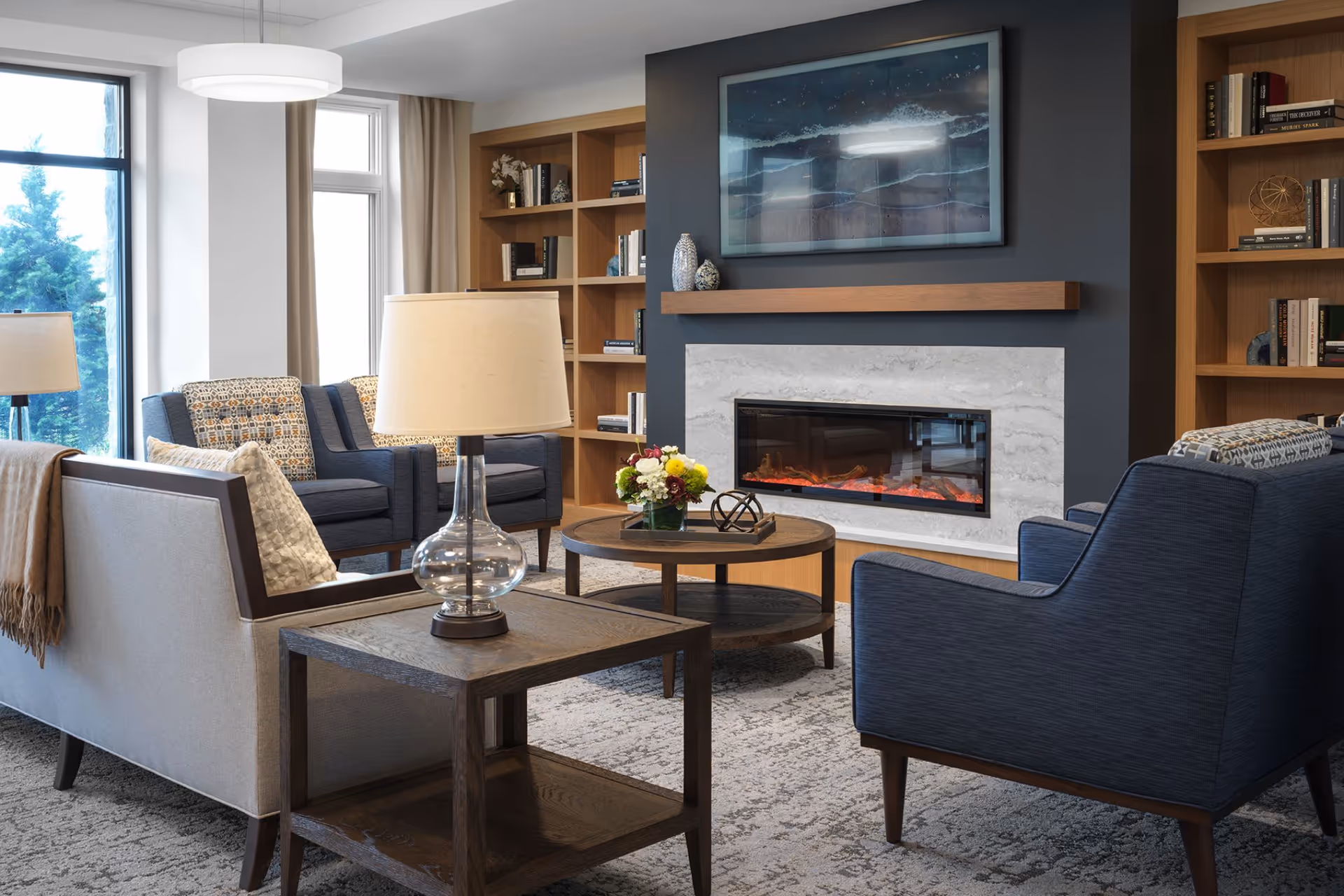 A cozy living room area with a modern electric fireplace set in a dark accent wall, flanked by wooden bookshelves filled with books and decorative items. The room features comfortable seating including a beige sofa with a throw blanket and pillows, two blue armchairs with patterned cushions, and a round wooden coffee table with a flower arrangement and decorative objects. A side table with a glass lamp is next to the sofa. Large windows with beige curtains allow natural light to fill the space.