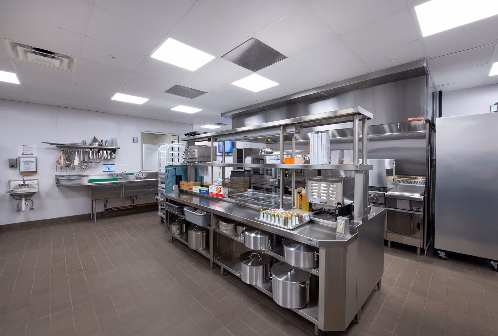 A clean, spacious commercial kitchen with stainless steel counters, shelves, and cooking equipment. There are pots and pans stored on the lower shelves, various kitchen utensils hanging on the wall, and a large refrigerator on the right side. The ceiling has bright fluorescent lights.