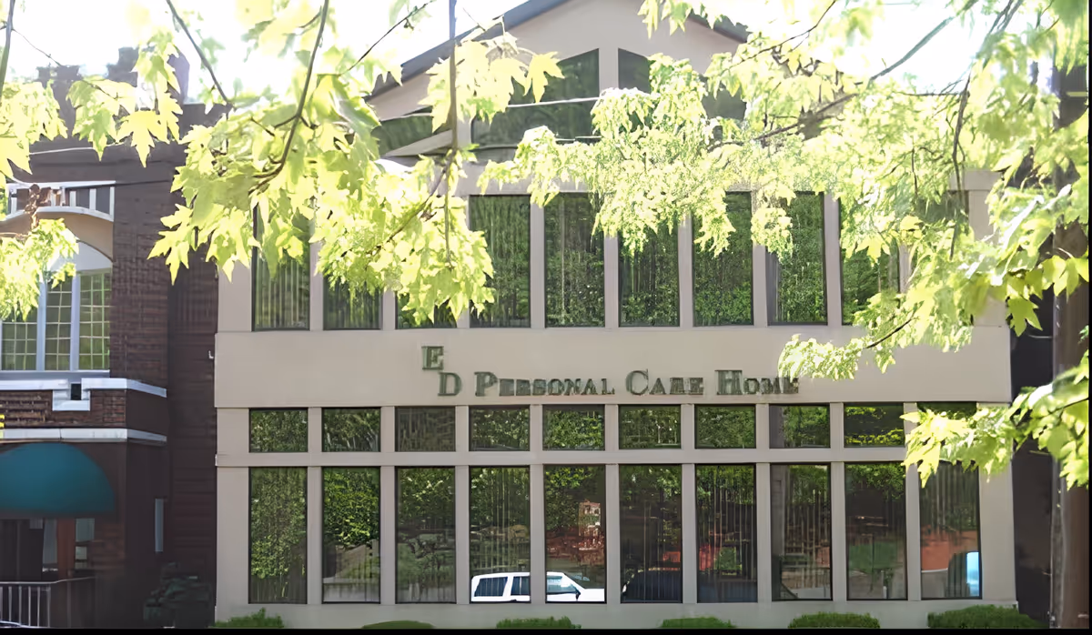 Exterior view of East Deer Personal Care Home building partially obscured by green tree branches with sunlight filtering through. The building has large windows reflecting the surrounding greenery and a white vehicle parked in front.