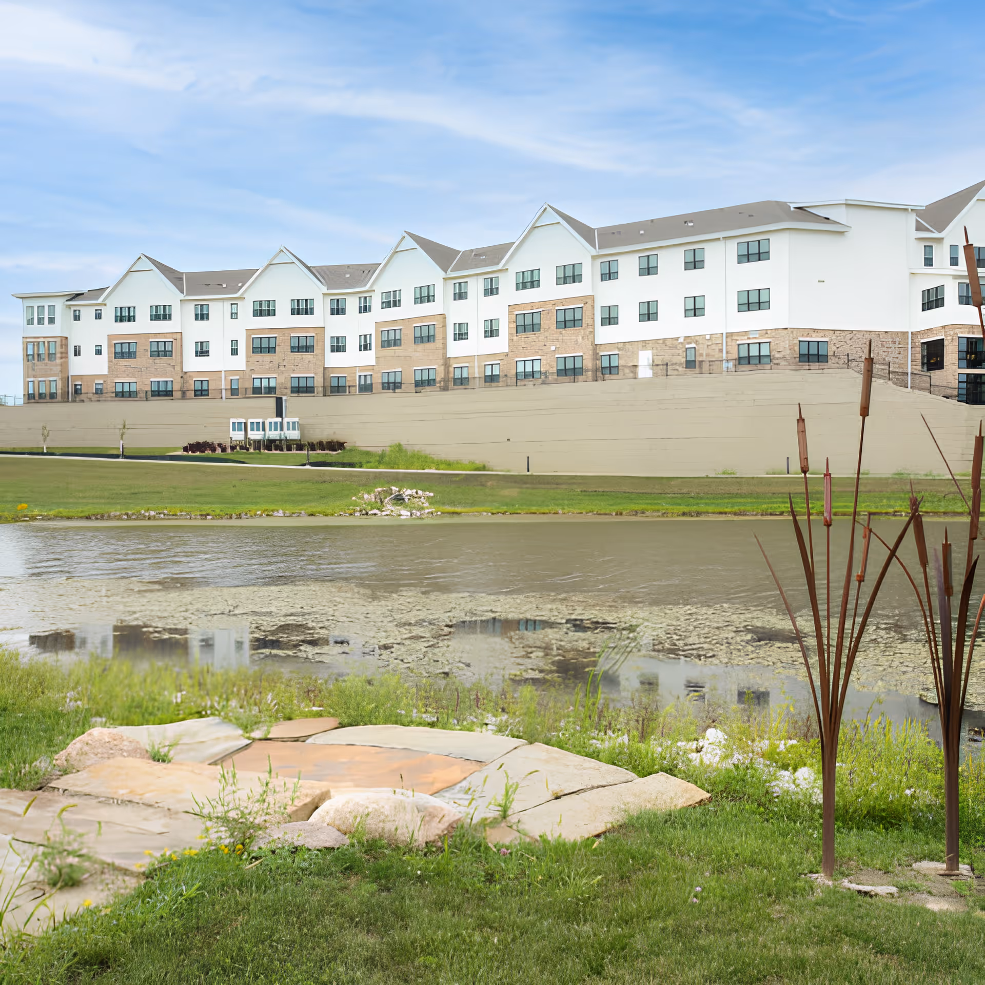 Large multi-story senior living building on a hill overlooking a pond and landscaped lawn with a stone patio in the foreground.