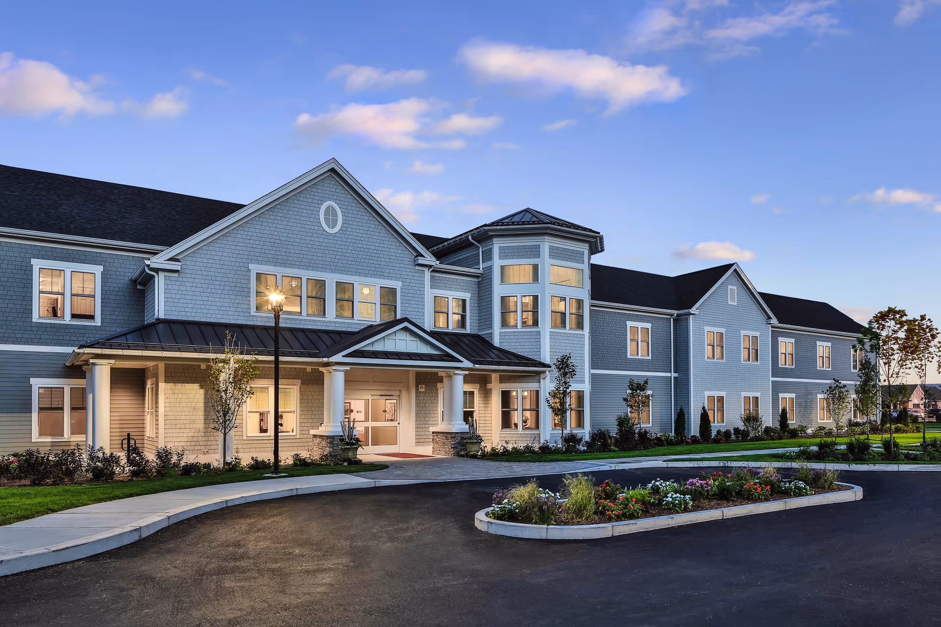 Blue-gray senior living building with a covered entrance, columns, and landscaped roundabout driveway.