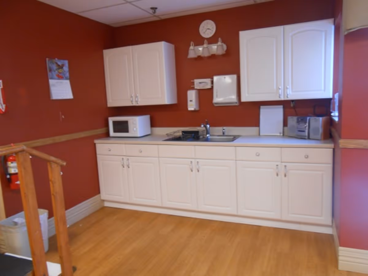 A small kitchenette area with white cabinets and drawers, a microwave on the counter, a sink, a clock and light fixture above, and a small stereo on the right side. The walls are painted dark red and the floor is light wood. A fire extinguisher is mounted on the left wall near a wooden railing.
