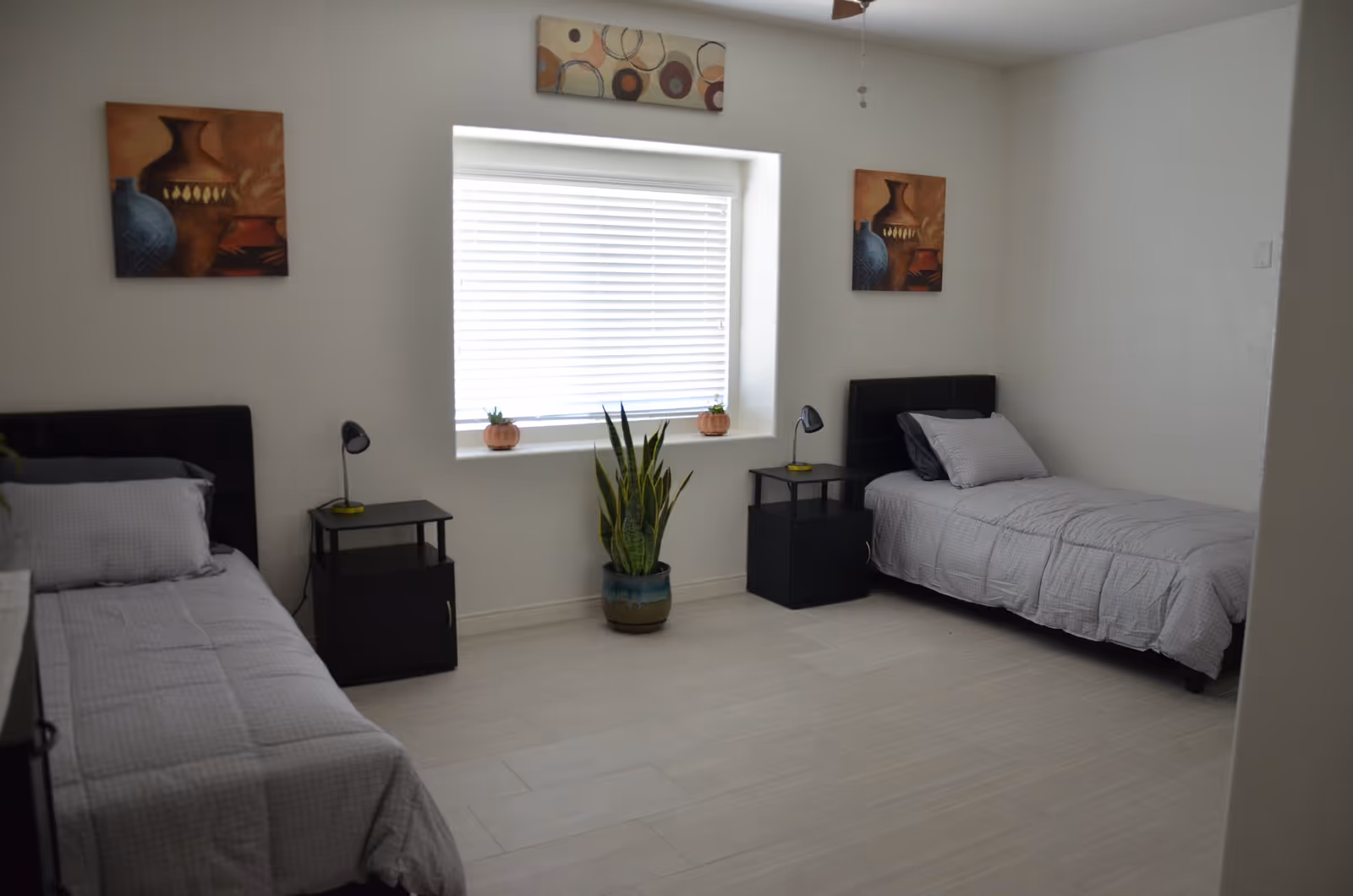 A simple bedroom with two single beds, nightstands, a potted plant, a window with blinds, and wall art.