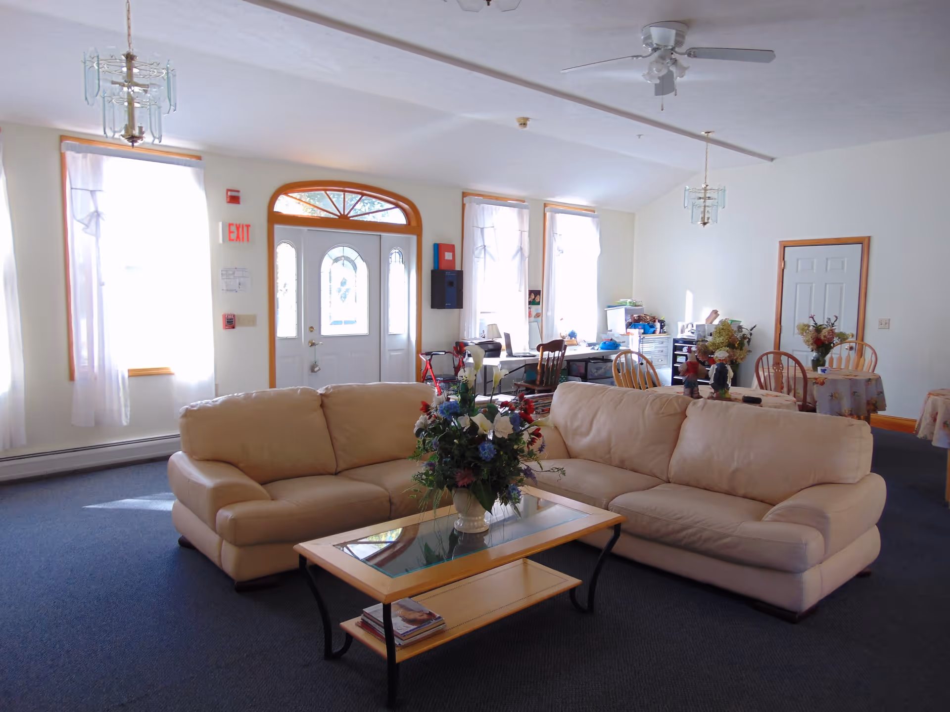 A bright communal living room with beige sofas, a glass coffee table topped with a floral arrangement, dining tables and chairs, and windows flanking an entrance door.