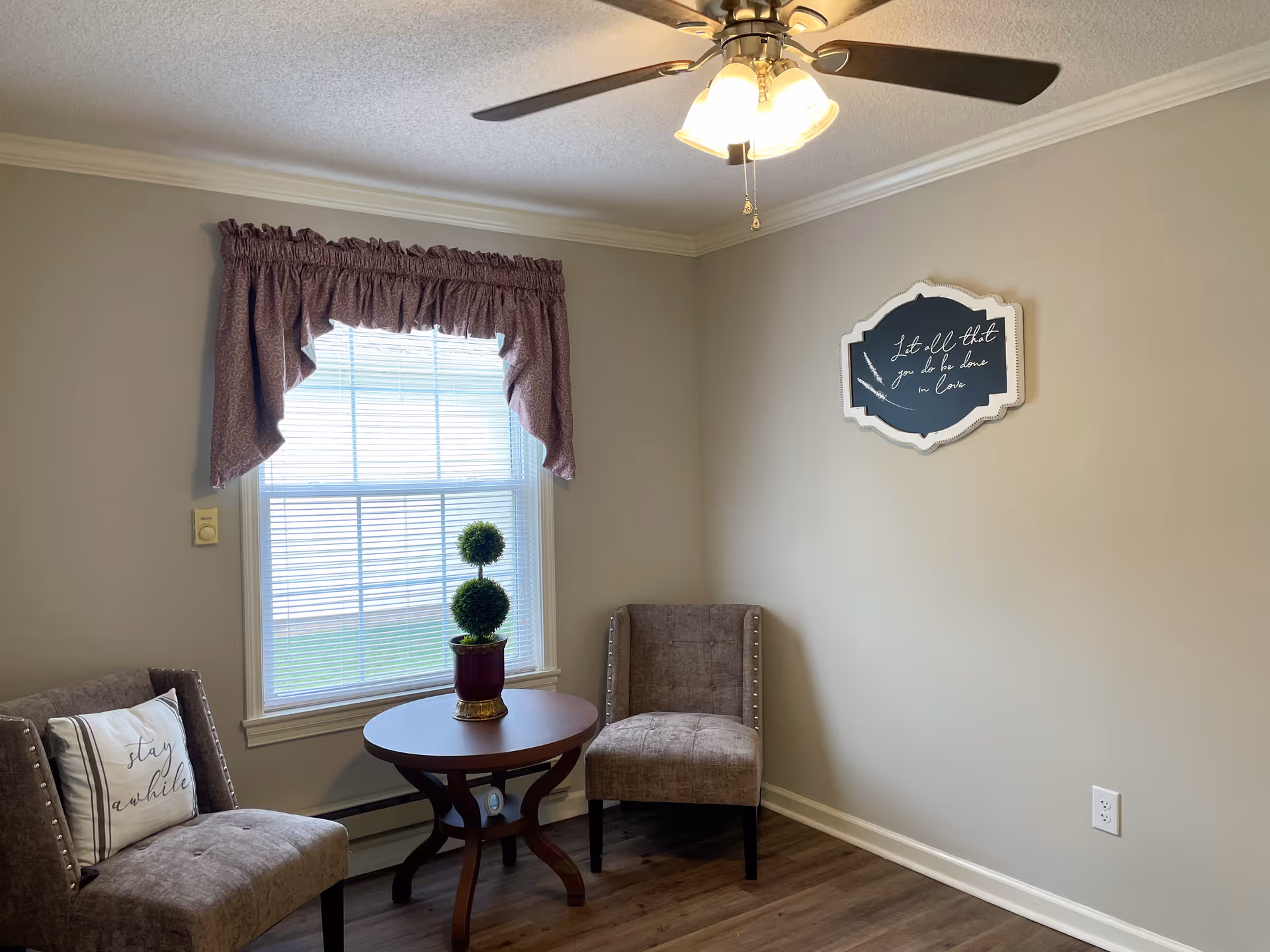 A small sitting area with two upholstered chairs and a round wooden table between them. A potted topiary plant sits on the table. There is a window with blinds and a purple valance curtain. A ceiling fan with a light is on the ceiling. A decorative wall sign with cursive writing is hung on the beige wall.