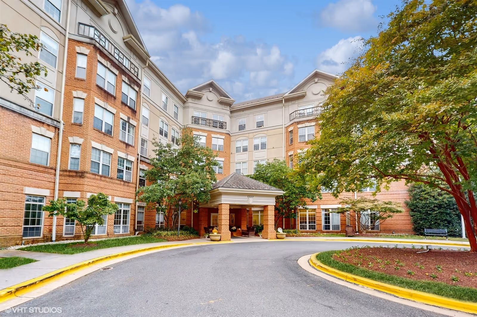 Front entrance of a multi-story brick senior living building with a covered porte-cochère and landscaped circular driveway.