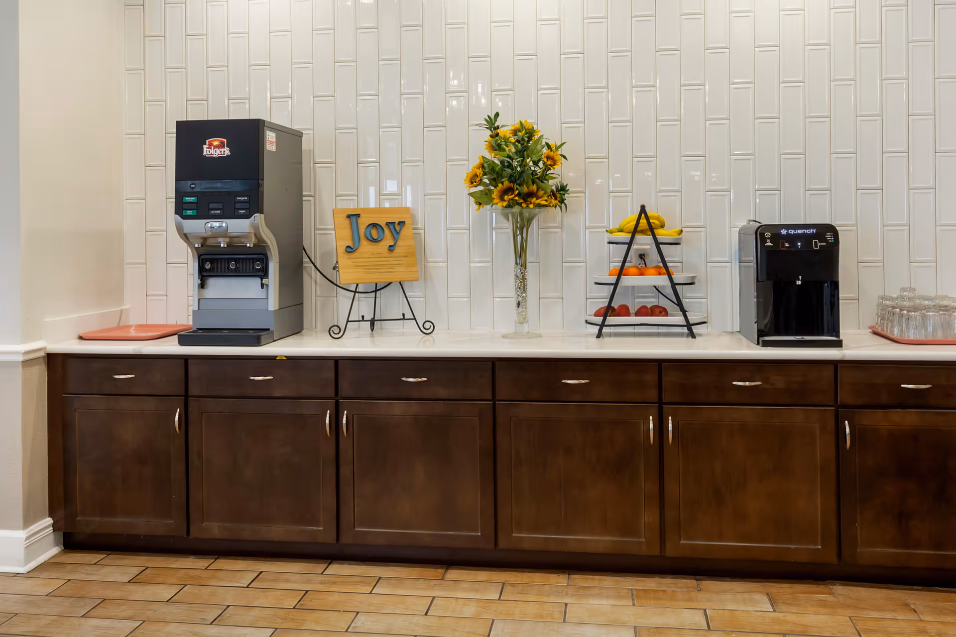 A countertop with a coffee machine on the left, a decorative sign that says 'Joy', a vase with sunflowers, a tiered fruit stand with bananas, oranges, and apples, and a water dispenser on the right. The background features a white tiled wall and dark wooden cabinets below the counter.