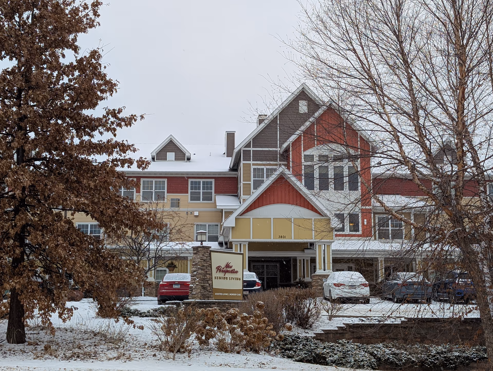 Exterior view of New Perspective Senior Living building in Columbia Heights during winter, showing a multi-story structure with red, beige, and brown siding, snow-covered ground, parked cars, and leafless trees.