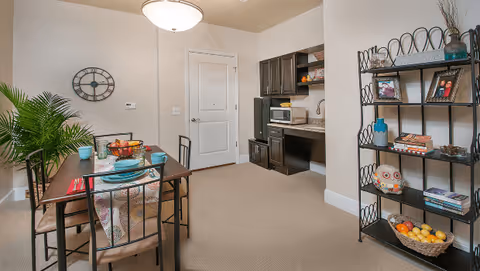 A dining area with a rectangular wooden table set for four with plates, bowls, and glasses. To the right, there is a black metal shelving unit holding books, framed photos, decorative items, and a basket of fruit. On the back wall, there is a kitchenette with dark cabinets, a microwave, and a small refrigerator. A large wall clock and a potted plant are visible on the left side of the image.