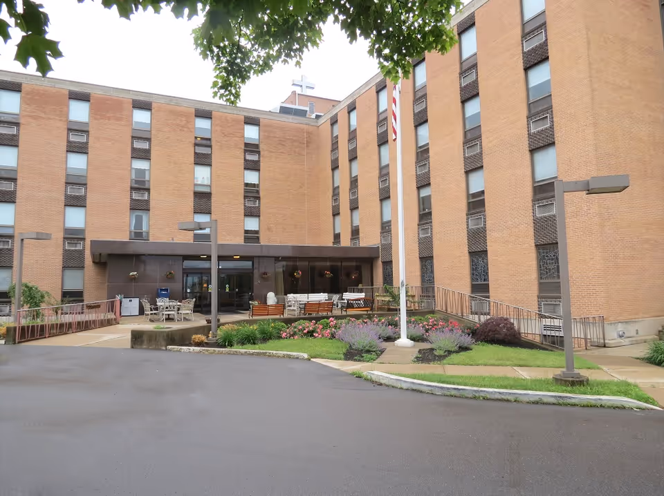Exterior view of a multi-story brick building with several windows, an entrance with glass doors, outdoor seating area with tables and chairs, a flagpole with an American flag, and landscaped flower beds in front. Trees partially frame the top of the image.