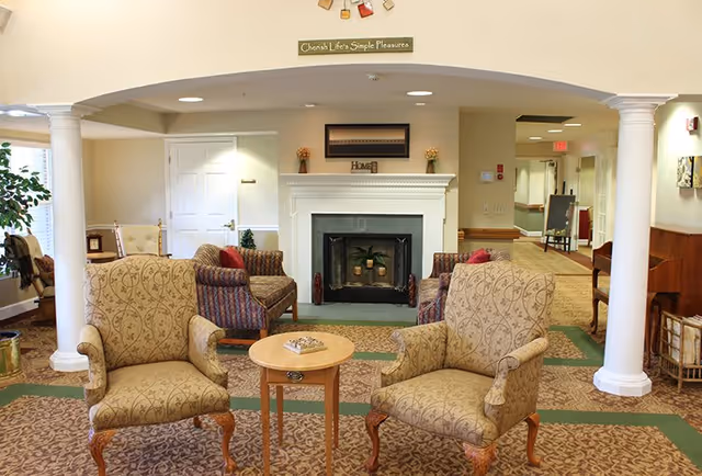 A cozy senior living community lounge area with two patterned armchairs and a small round wooden table in the foreground. Behind them are two more armchairs with striped upholstery facing a white fireplace with decorative items on the mantel. The room features beige walls, carpeted floors, white columns, and a sign above the fireplace that reads 'Cherish Life's Simple Pleasures.'