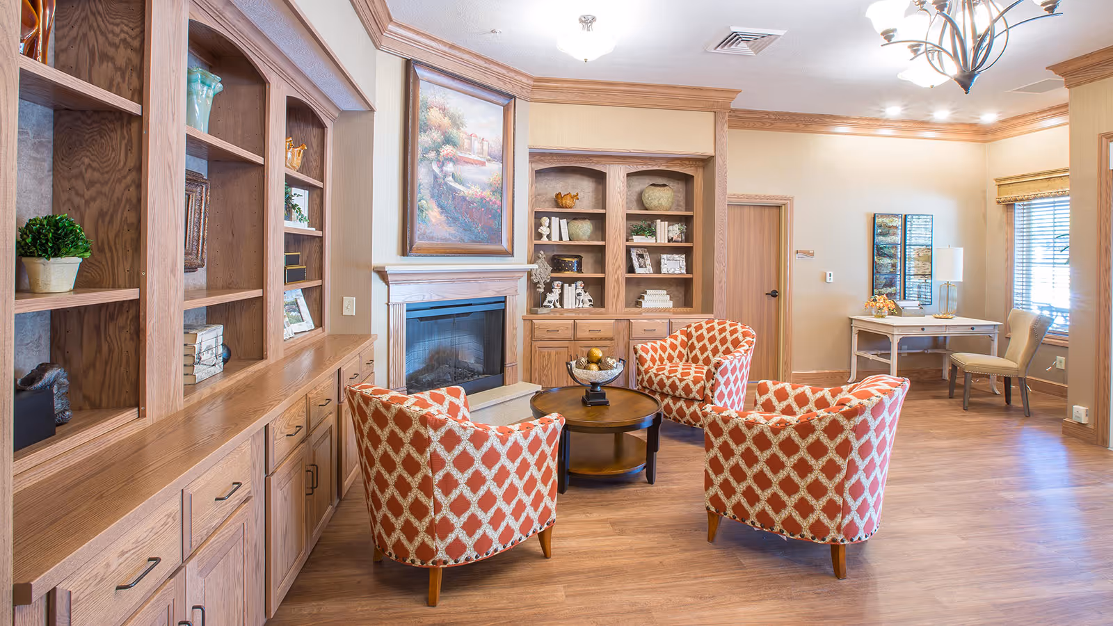 A cozy living room area in a senior living facility featuring three orange and white patterned armchairs arranged around a round wooden coffee table. The room has wooden built-in shelves and cabinets, a fireplace with a large framed painting above it, a desk with a chair near a window with blinds, and warm lighting from ceiling fixtures.