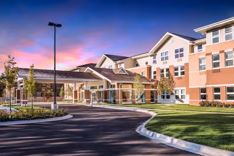 Exterior view of a large, modern senior living facility building with a covered entrance, surrounded by landscaped greenery and a curved driveway under a colorful sunset sky.