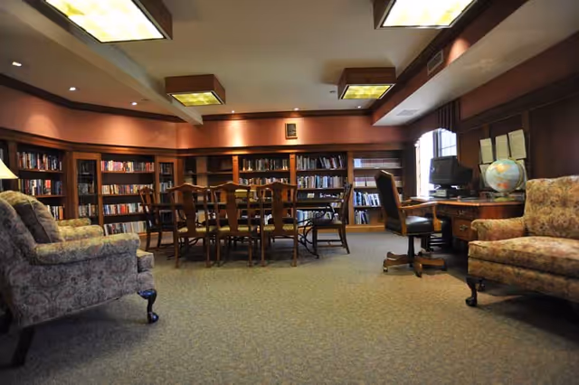Cozy senior living library lounge with bookshelves, tables, chairs, a computer desk, and globe.