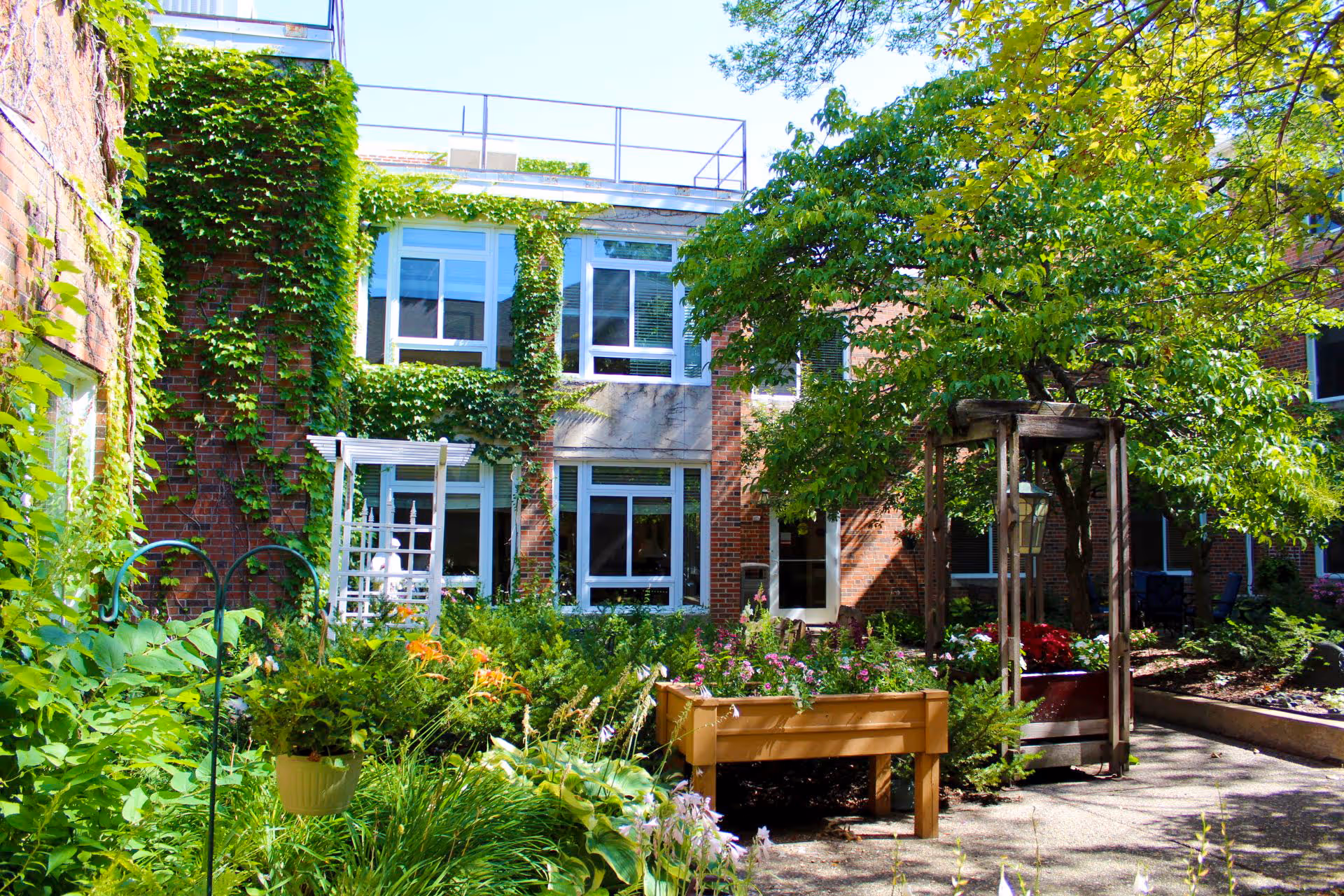 A bright outdoor garden area at Episcopal Homes of Minnesota featuring lush green plants, flowers, a small tree supported by a wooden frame, and a brick building covered partially with ivy in the background.