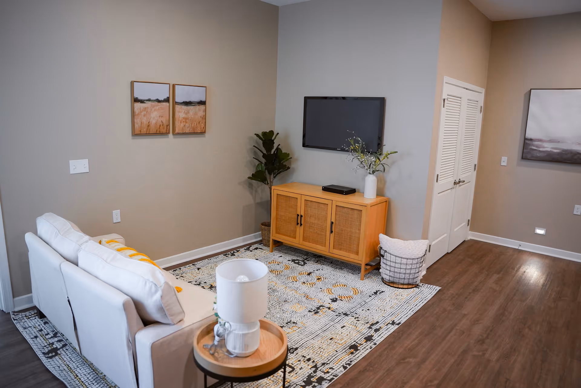 A cozy living room with a white sofa, a patterned area rug, a wooden side table with a white lamp, a wooden cabinet with a vase of flowers, a flat-screen TV mounted on the wall, two landscape paintings, a potted plant, and a basket with a pillow on a wooden floor.