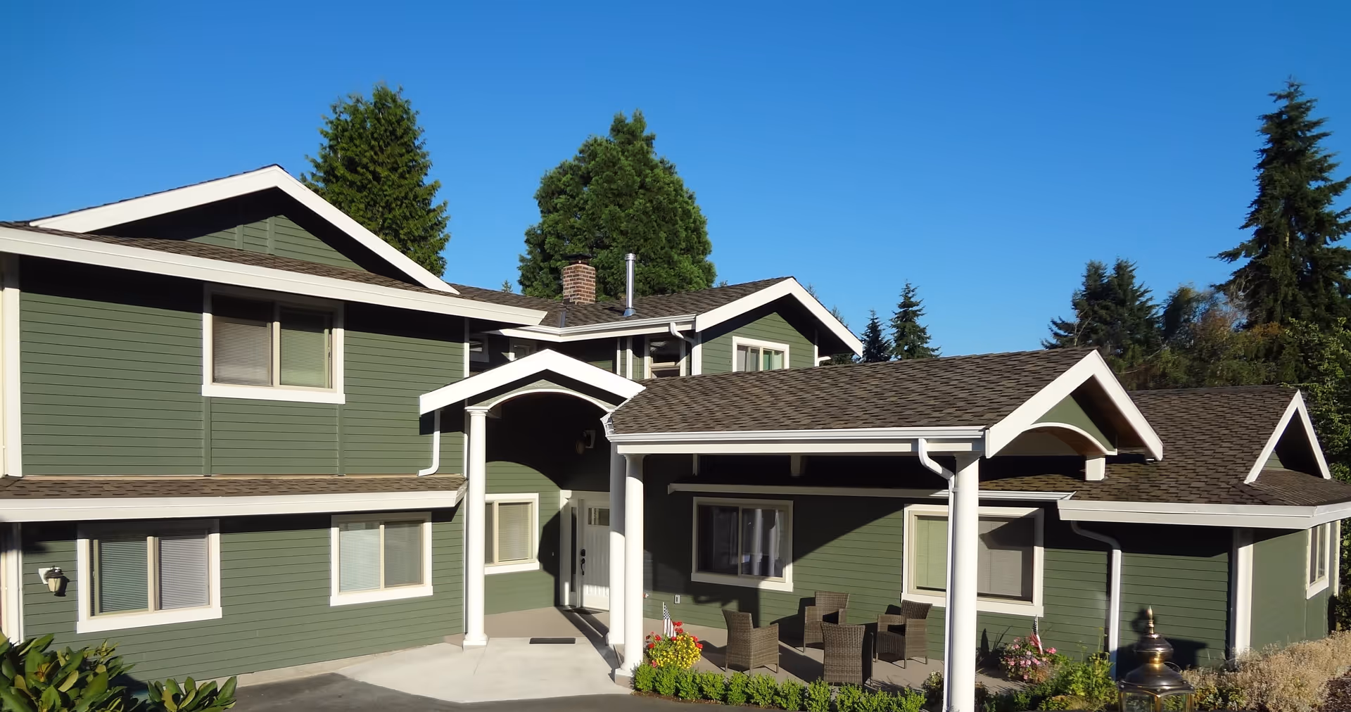 Exterior view of a green two-story building with white trim and a covered entrance. There are outdoor chairs and a small table on the porch, surrounded by plants and trees under a clear blue sky.