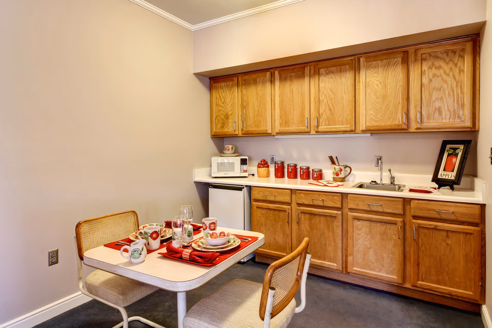 Small kitchen area with wooden cabinets, a white countertop, a microwave, a small refrigerator, and a sink. A table set for two with apple-themed dishes, red napkins, and glassware is positioned in front of the cabinets. The walls are beige and the floor is dark-colored.