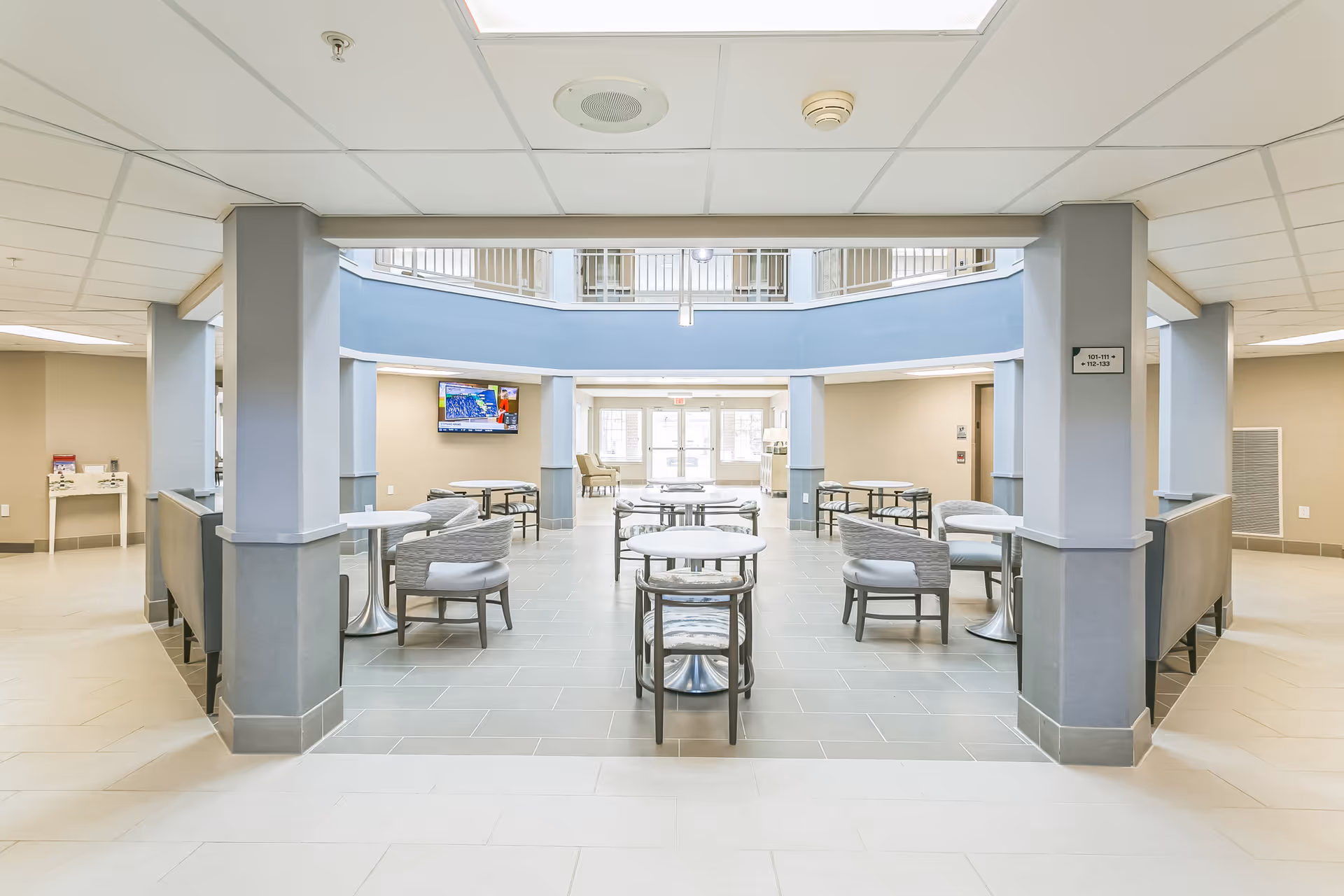 Bright open senior living common area with round tables and chairs beneath a second-floor atrium.