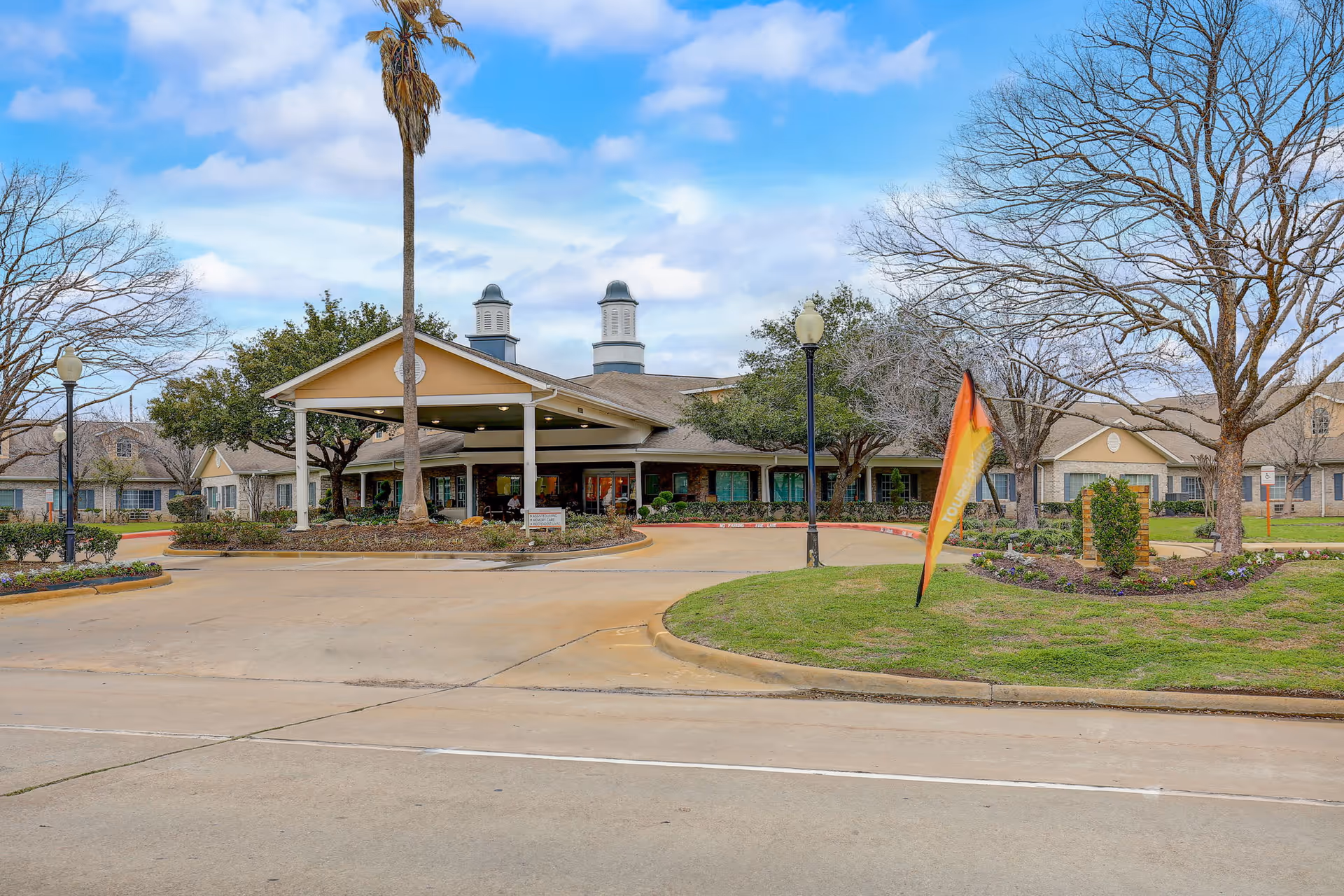 Front exterior view of Morada Cy-Fair senior living facility with a covered entrance, palm tree, lamp posts, and landscaped greenery under a partly cloudy sky.